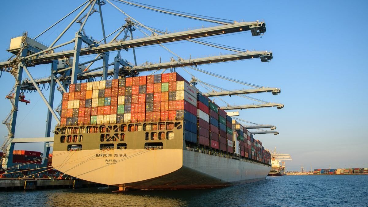 A large container ship at an industrial harbor with cranes under a clear blue sky