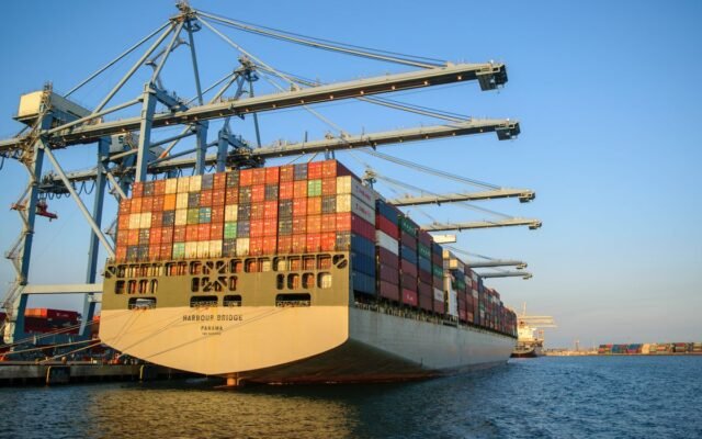 A large container ship at an industrial harbor with cranes under a clear blue sky