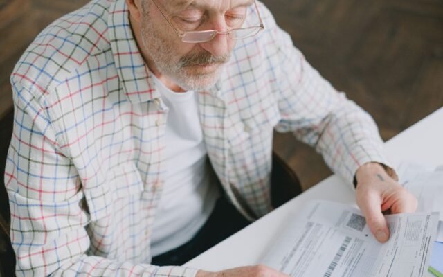Elderly man in glasses carefully examines documents at home. Thoughtful and focused