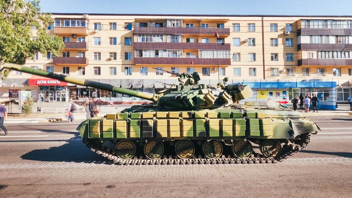 Camouflaged military tank parked in front of an urban residential building during a sunny day
