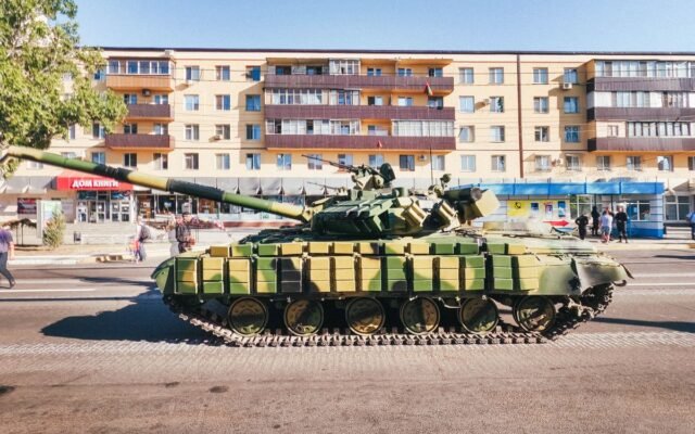 Camouflaged military tank parked in front of an urban residential building during a sunny day